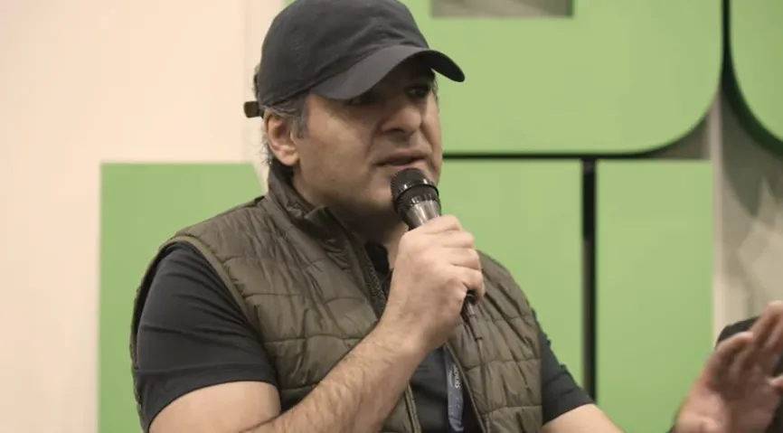 A man wearing a cap and vest speaking into a microphone during an event.
